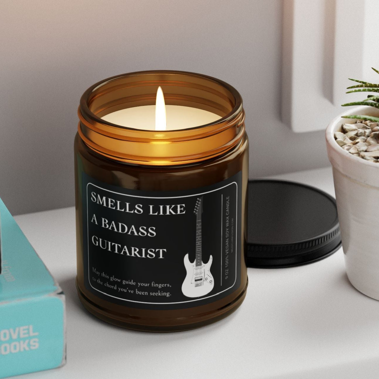 Candle in an amber jar labeled 'Smells Like a Badass Guitarist' on a shelf with books and a plant.