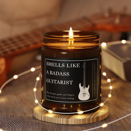 Candle in a jar labeled 'Smells Like a Badass Guitarist' with a guitar on a wooden surface.