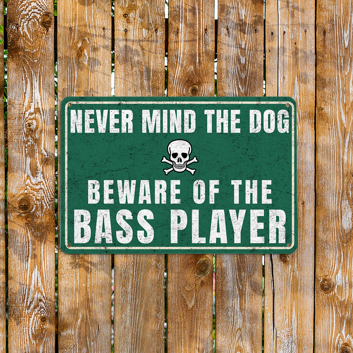 A vintage-style green metal sign with a skull and crossbones icon that reads 'Never mind the dog, beware of the bass player' mounted on a rustic wooden fence.