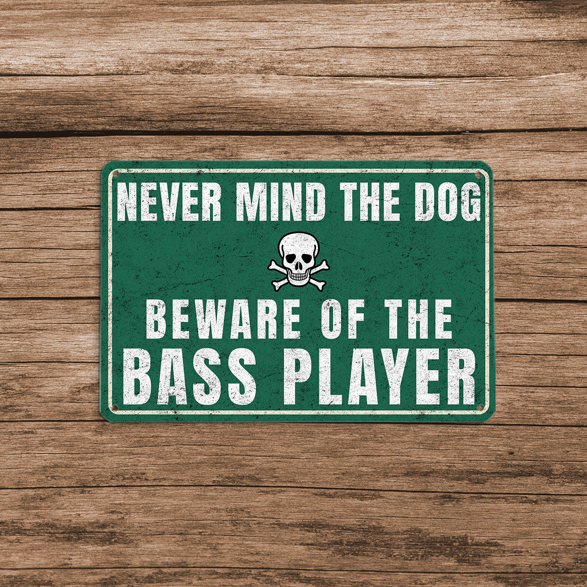 A vintage-style green metal sign with a skull and crossbones icon that reads 'Never mind the dog, beware of the bass player' mounted on a rustic wooden fence.