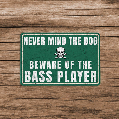A vintage-style green metal sign with a skull and crossbones icon that reads 'Never mind the dog, beware of the bass player' mounted on a rustic wooden fence.