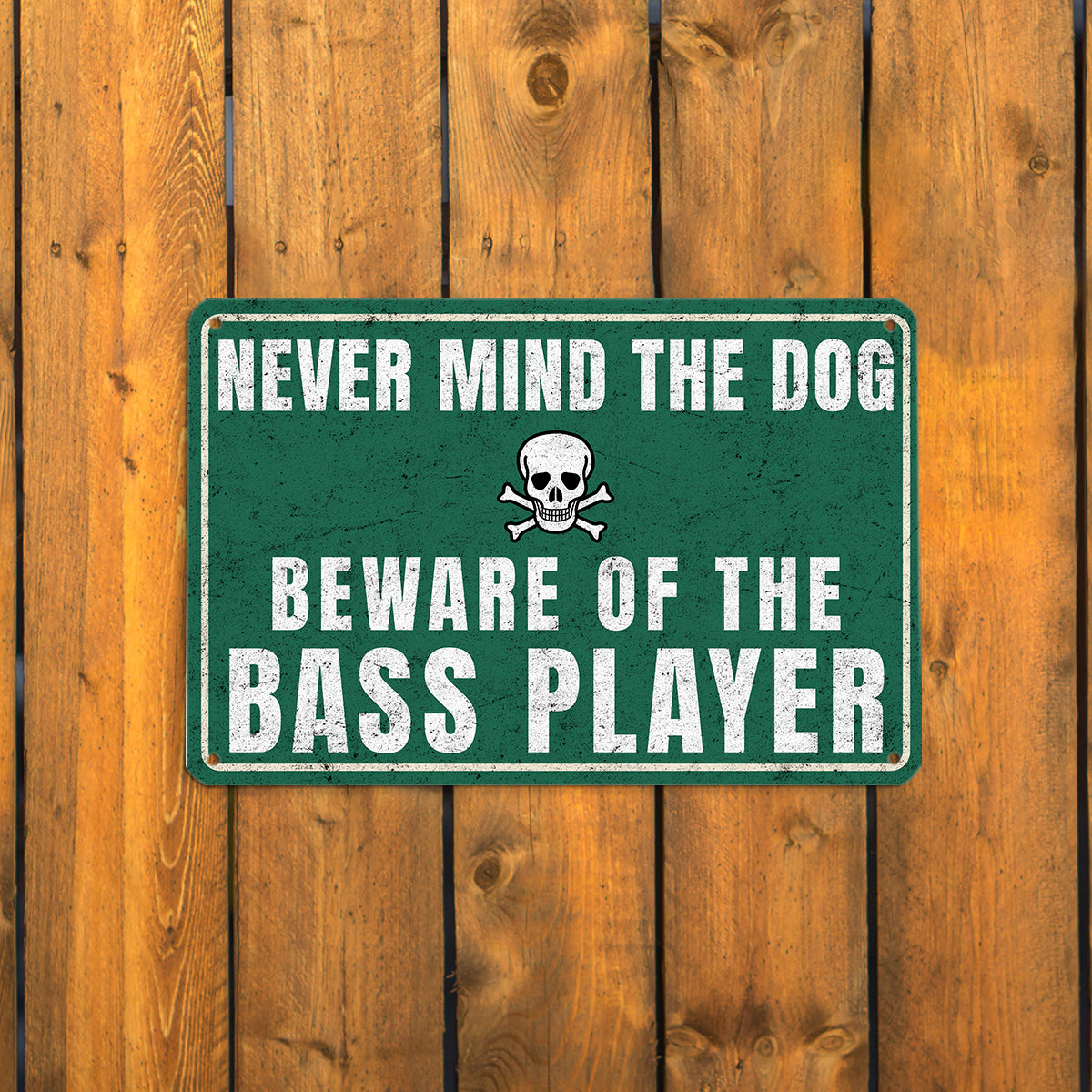 A vintage-style green metal sign with a skull and crossbones icon that reads 'Never mind the dog, beware of the bass player' mounted on a rustic wooden fence.