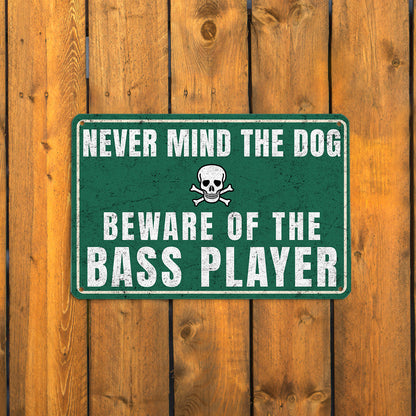 A vintage-style green metal sign with a skull and crossbones icon that reads 'Never mind the dog, beware of the bass player' mounted on a rustic wooden fence.