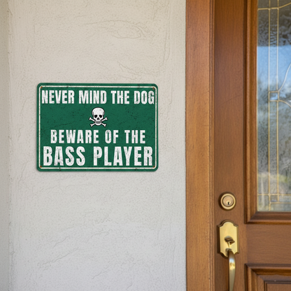A vintage-style green metal sign with a skull and crossbones icon that reads 'Never mind the dog, beware of the bass player' mounted on a rustic wooden fence.