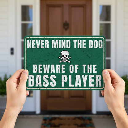 A vintage-style green metal sign with a skull and crossbones icon that reads 'Never mind the dog, beware of the bass player' mounted on a rustic wooden fence.