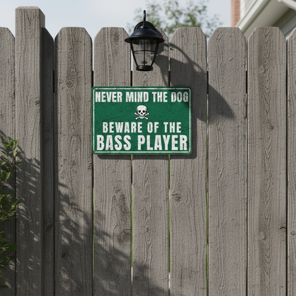 A vintage-style green metal sign with a skull and crossbones icon that reads 'Never mind the dog, beware of the bass player' mounted on a rustic wooden fence.